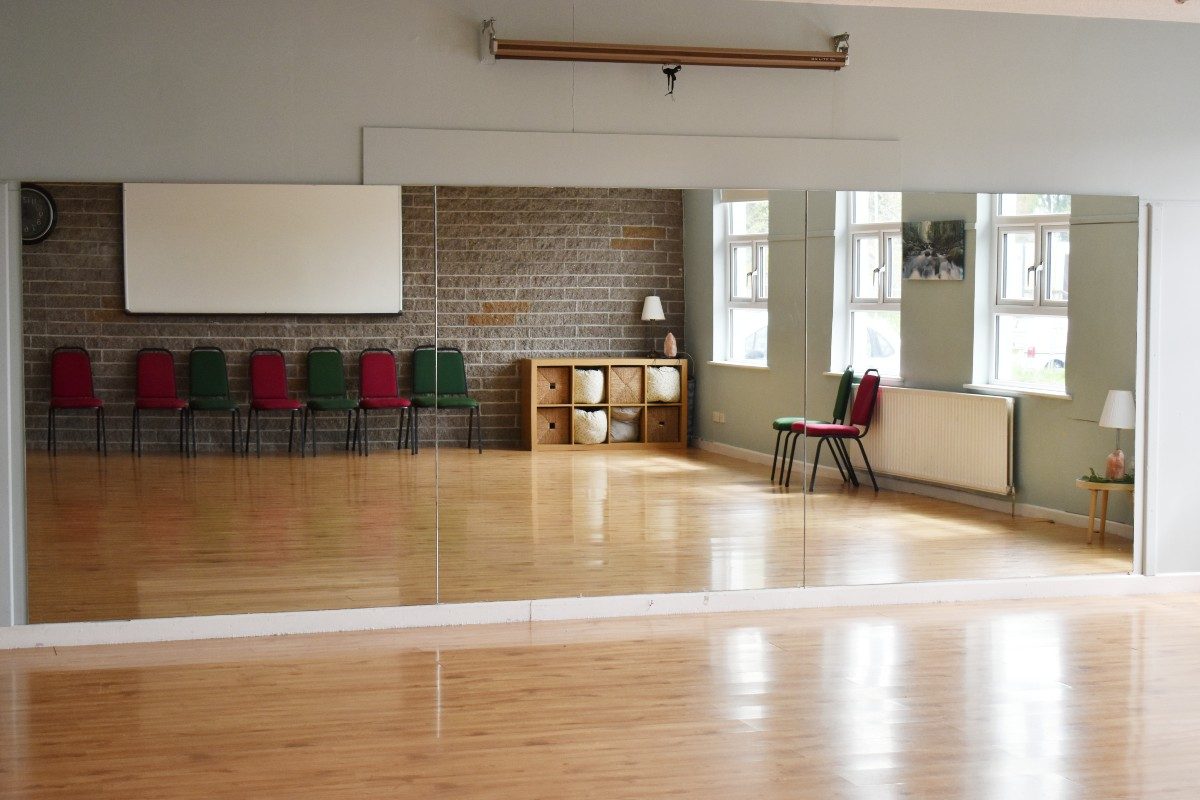 Physical activity room with mirrors, wooden floor, and green and red chairs.