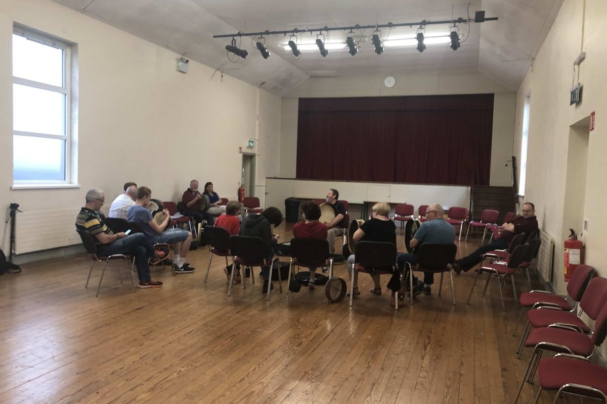 St. James' Hall: Musicians playing instruments in a circle, seated on chairs in the hall.