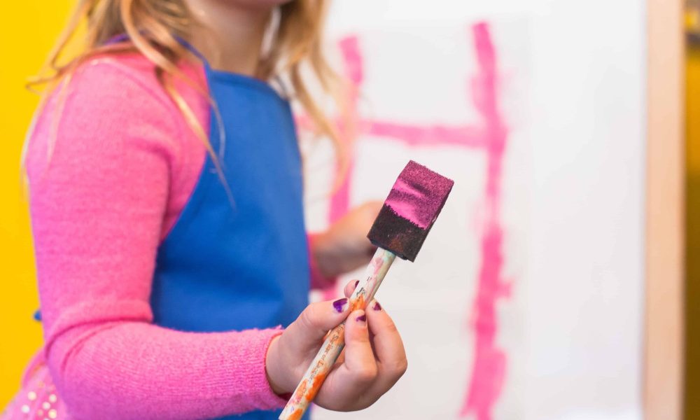 A young girl with a paintbrush in hand, standing in front of a white board, ready to create art.